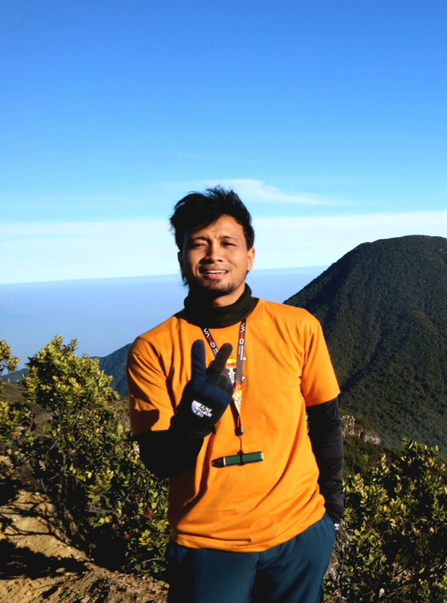 Man wearing orange t-shirt with black gloves standing in front of mountain and blue sky