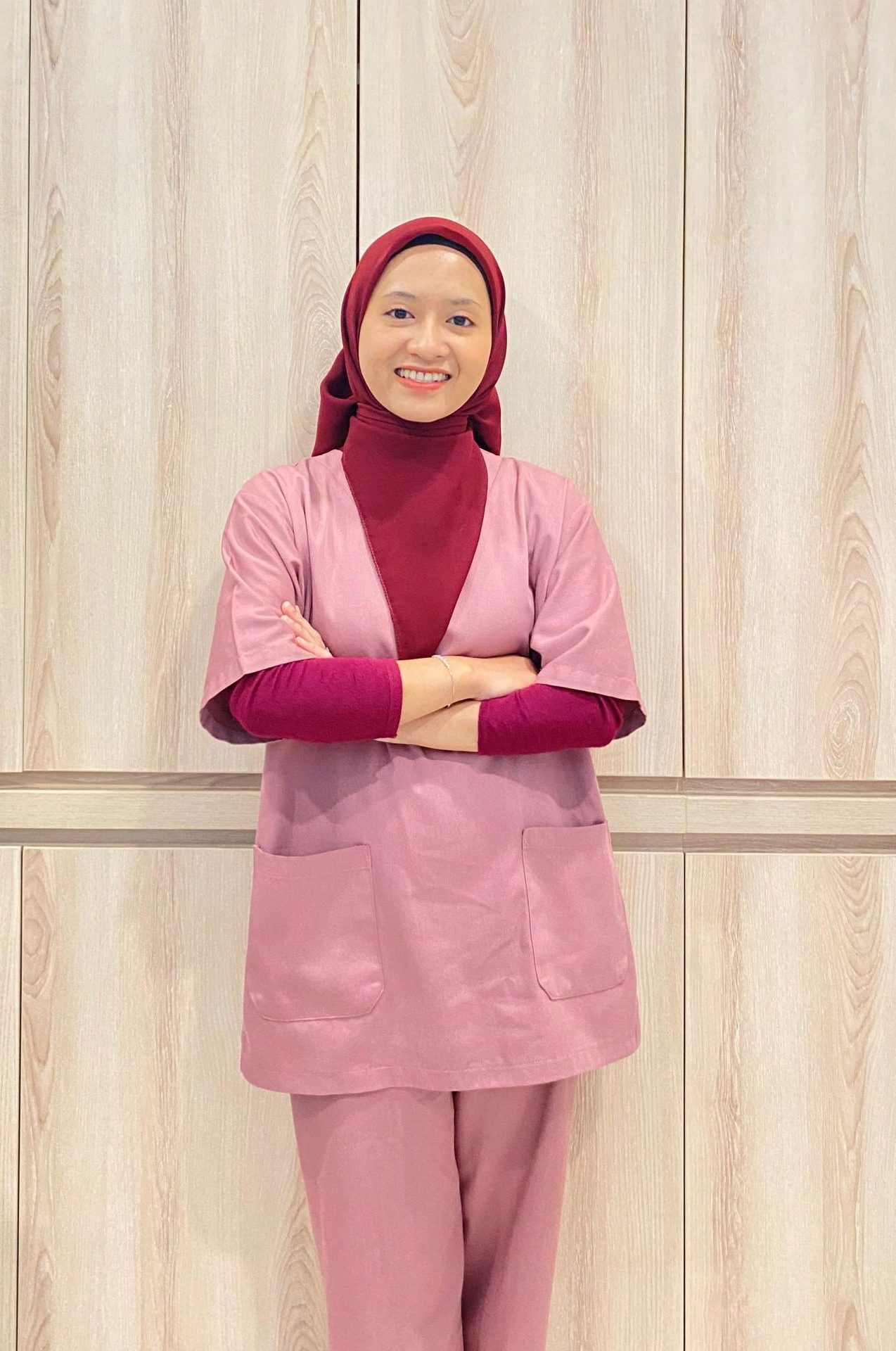 Woman wearing red hijab and pink outerwear smiling with arms crossed in front of wooden wall